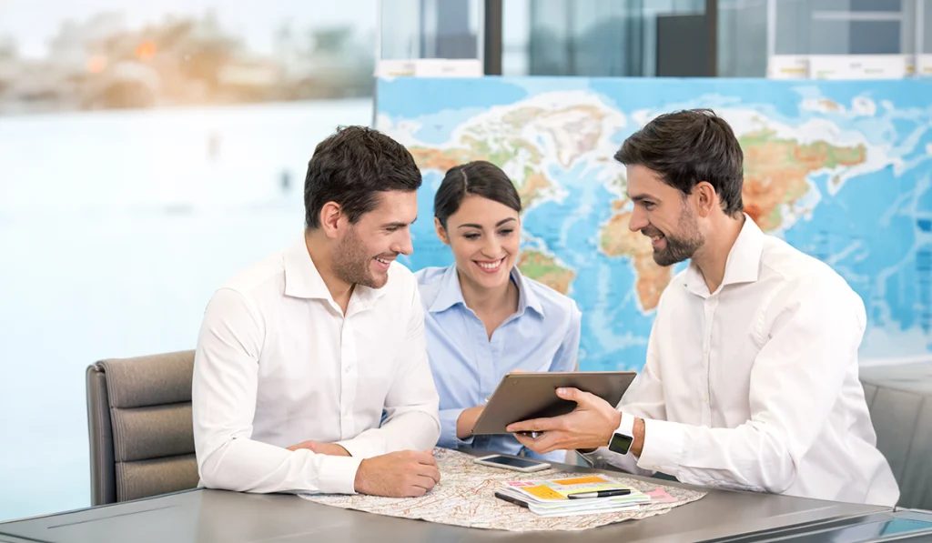 Travel company travel agents assisting clients with personalized holiday planning using a tablet, maps, and travel brochures in a professional travel agency office.